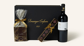 Das Bild zeigt ein elegantes Geschenkset aus der Breuninger Confiserie. Im Zentrum steht eine schwarze Box mit goldener Aufschrift „Breuninger Confiserie 1951“. Neben der Box befinden sich drei hochwertige Produkte:  Links ist ein transparent verpackte Schokoladentafel, mit einer goldenen Schleife verziert. In der Mitte liegt eine längliche Schachtel mit Pralinen, ebenfalls mit einer goldenen Schleife umwickelt. Rechts steht eine Flasche Merlot mit einem weißen Etikett. Das Set vermittelt einen luxuriösen und stilvollen Eindruck, ideal als Geschenk für besondere Anlässe.