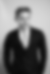 Schwarz-weiß Porträt eines Mannes im Anzug. Das Bild ist stark unscharf, sodass Gesichtszüge und Details nicht erkennbar sind. Der Mann steht mit den Händen in den Hosentaschen vor neutralem Hintergrund.