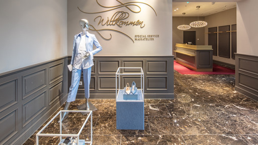 Eingangsbereich eines Modegeschäfts mit Schaufensterpuppe, Präsentationspodesten, Marmorboden und Empfangstheke im Hintergrund.