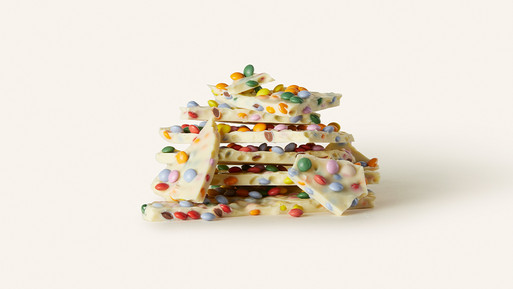 Gestapelte Bruchstücke weißer Schokolade mit bunten Zuckerdragées auf hellem Hintergrund.