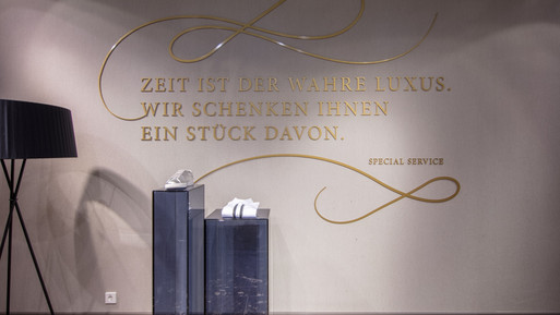 Wandgestaltung mit goldener Schrift „Zeit ist der wahre Luxus. Wir schenken Ihnen ein Stück davon.“ sowie Präsentationspodesten in einem eleganten Innenraum.