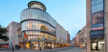 Außenansicht eines mehrstöckigen Breuninger-Kaufhauses mit abgerundeter Glasfassade an einer Straßenecke; beleuchtete Schaufenster im Erdgeschoss, umliegende Stadtbebauung und Gehweg im Vordergrund.