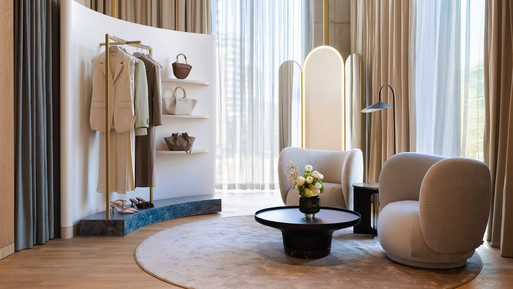 Heller Lounge-Bereich mit rundem Teppich, zwei gepolsterten Sesseln und niedrigem Couchtisch. Dahinter ein freistehender Spiegel und eine Kleiderstange mit hellen Kleidungsstücken. Warme Holzelemente, bodentiefe Vorhänge und sanfte Beleuchtung erzeugen eine ruhige, elegante Atmosphäre.