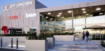 Außenansicht eines modernen Einkaufszentrums mit breitem Eingangsbereich. Vor dem Gebäude stehen Poller und Sitzmöglichkeiten, mehrere Personen betreten oder verlassen das Center. Die Fassade ist hell und großflächig verglast.