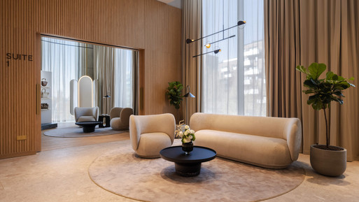 Moderner Innenraum mit hellem Lounge-Sofa, rundem Teppich und niedrigem Couchtisch in der Mitte. Warme Holzwände, bodentiefe Fenster mit Vorhängen und dezente Pflanzen schaffen eine ruhige, elegante Atmosphäre.