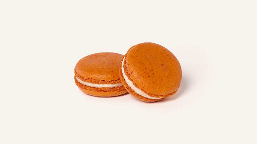 Zwei orangefarbene Pumpkin-Spice-Macarons mit heller Cremefüllung, dekoriert mit einem feinen weißen Schokoladenstreifen auf der Oberseite, vor hellem Hintergrund.