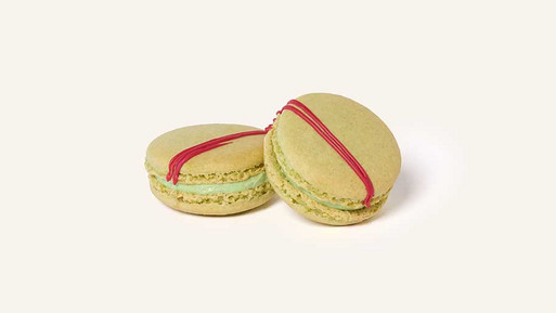 Zwei hellgrüne Matcha-Strawberry-Macarons mit zarter, grünlicher Füllung, dekoriert mit einem diagonalen Streifen aus roter Glasur auf der Oberseite, vor hellem Hintergrund.