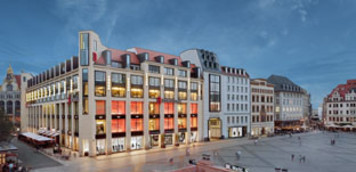 Mehrstöckiges Kaufhausgebäude mit moderner Fassade und großflächigen Schaufenstern, abends beleuchtet, an einer Straßenecke in der Innenstadt