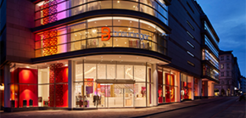 Fassade des Breuninger Flagship‑Stores in München am Abend, verglaster Eingangsbereich mit rotem Markenlogo und beleuchteten Schaufenstern