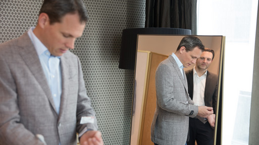 Mann probiert ein Sakko an, während ein weiterer Mann ihn im Spiegel in einem eleganten Beratungsbereich betrachtet.