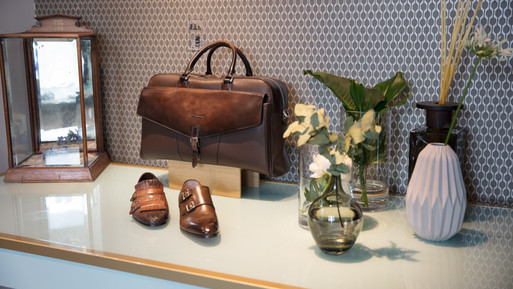 Präsentation mit Ledertasche, Herrenschuhen und dekorativen Vasen auf einer Ablage vor gemusterter Wand.
