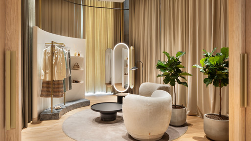 Eleganter Beratungsraum mit rundem Teppich, niedrigen Polstersesseln und einem kleinen schwarzen Beistelltisch in der Mitte. Helle Holzpaneele, bodentiefe Vorhänge und warme, indirekte Beleuchtung erzeugen eine ruhige Atmosphäre. Links ein Kleiderständer mit ausgewählten Kleidungsstücken, rechts große Pflanzen in schlichten Gefäßen sowie ein freistehender Spiegel.