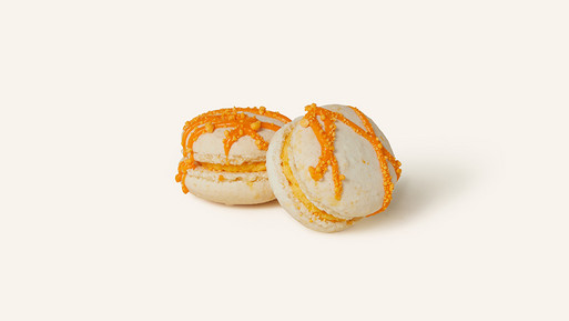 Zwei Champagner-Orange-Macarons mit einer zart-cremefarbenen Schale, dekoriert mit leuchtend orangefarbenen Streifen. Gefüllt mit einer aromatischen Ganache, vereinen sie die spritzige Frische von Orange mit der Eleganz von Champagner – ein luxuriöser Genuss aus der Breuninger Confiserie.