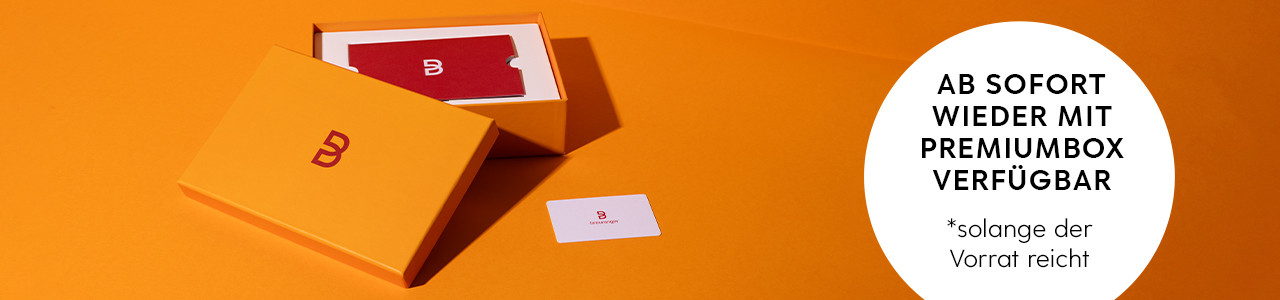 Orange Box mit Breuninger-Logo, rote und weiße Karte, Text: „Ab sofort wieder mit Premiumbox verfügbar“.