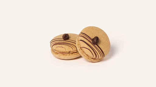 Zwei beige Espresso-Martini-Macarons mit heller Füllung, dekoriert mit feinen dunklen Schokoladenlinien und jeweils einer ganzen Kaffeebohne auf der Oberseite, vor hellem Hintergrund.