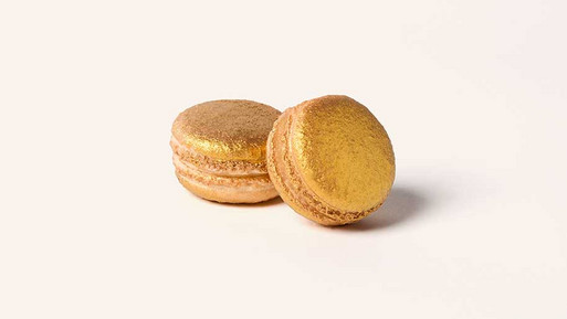 Zwei Bratapfel-Macarons in warmem Beigeton mit heller Füllung, deren Oberseiten vollständig mit goldfarbener Dekoration überzogen sind, vor hellem Hintergrund.