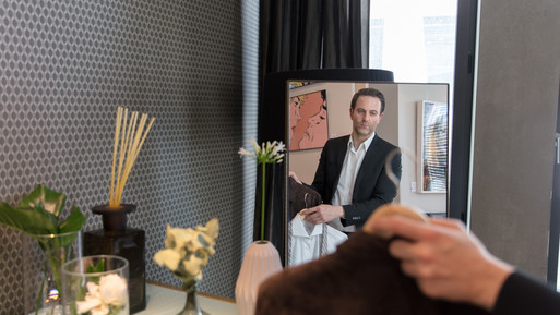 Mann betrachtet ein Kleidungsstück vor einem Spiegel in einem stilvoll eingerichteten Beratungs- oder Ankleidebereich.