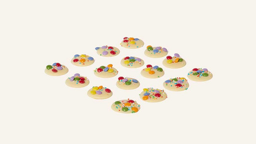 Viele kleine, runde Palets au Chocolat aus weißer Schokolade mit bunten Schokolinsen in Rasteranordnung auf hellem Hintergrund – feine, französische Schokoladenplätzchen mit farbenfrohem, spielerischem Finish aus der Breuninger Confiserie.