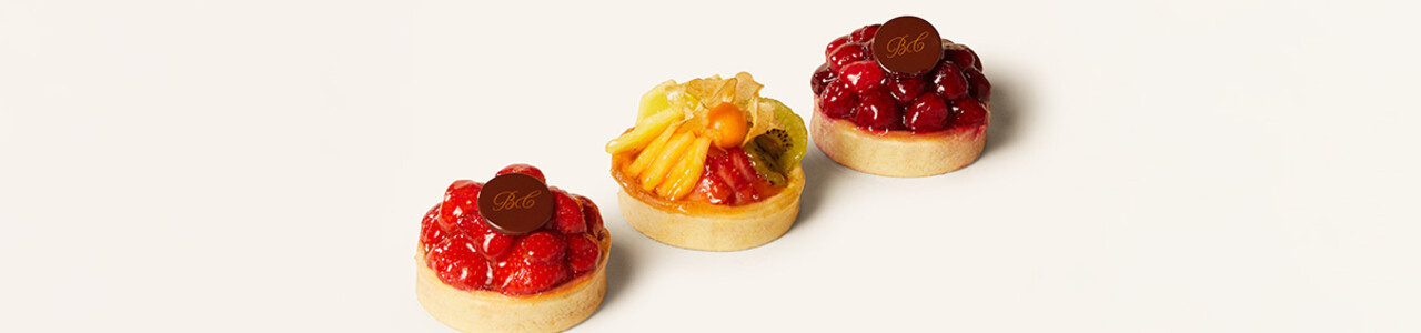 Drei elegante Fruchttörtchen in filigranen Mürbeteigböden, jeweils kunstvoll mit frischen Früchten belegt. Das erste und dritte Törtchen sind mit glänzenden Erdbeeren bedeckt, das mittlere Törtchen ist mit einer Mischung aus exotischen Früchten wie Kiwi, Mango und Physalis dekoriert. Jedes Törtchen ist mit einem kleinen Schokoladenemblem versehen, das ein stilvolles „BC“ trägt. Die Törtchen wirken hochwertig und einladend.