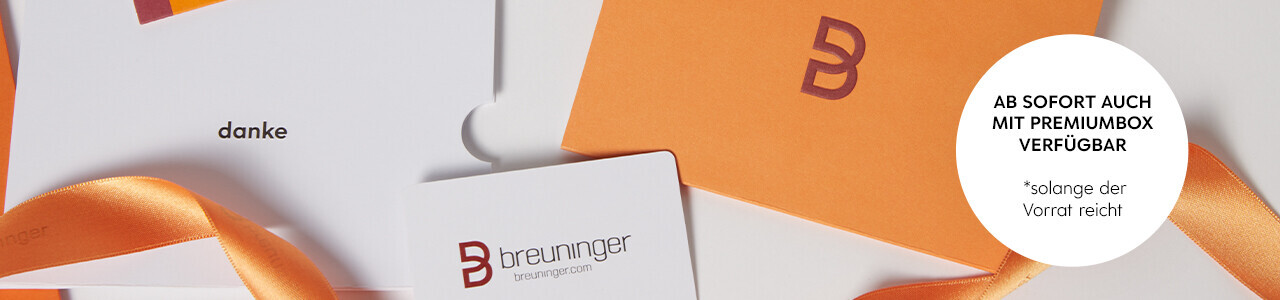 Abgebildet ist eine Breuninger-Geschenkkarte in Weiß mit rotem Logo, zusammen mit einer orangefarbenen Premium-Geschenkverpackung und passendem Satinband. Ein Kärtchen mit dem Aufdruck ‚danke‘ ist ebenfalls zu sehen. Rechts im Bild befindet sich ein weißer Kreis mit dem Hinweis: ‚Ab sofort auch mit Premiumbox verfügbar – solange der Vorrat reicht