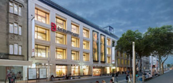 Außenansicht des Breuninger Hauses in Karlsruhe in den Abendstunden. Die Fassade ist hell beleuchtet, das rote Breuninger-Logo prangt gut sichtbar an der oberen Ecke. Vor dem Gebäude befinden sich Bäume, Straßenlaternen und Passanten.