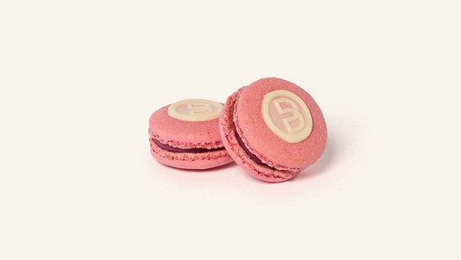 Zwei rosafarbene Sommerbeeren-Macarons mit dunkler Füllung und einem runden Siegel aus weißer Schokolade mit Breuninger „B“ auf der Oberseite, vor hellem Hintergrund