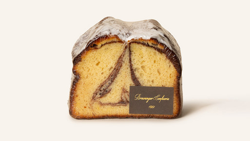 Ein saftiger Marmorkuchen, der goldbraun gebacken ist. Er hat kunstvolle Schokoladenwirbel im Inneren, der den perfekten Genussmoment verspricht. Der Kuchen ist mit Puderzucker bedeckt und trägt ein kleines, edles Schild mit der Aufschrift "Confiserie Breuninger". Ein Klassiker, der Tradition und Qualität in jedem Bissen vereint.