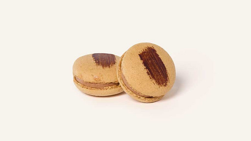 Zwei hellbraune Café-Macarons mit dunkler Cremefüllung, dekoriert mit einem breiten Pinselstrich aus dunkler Schokolade auf der Oberseite, vor hellem Hintergrund.