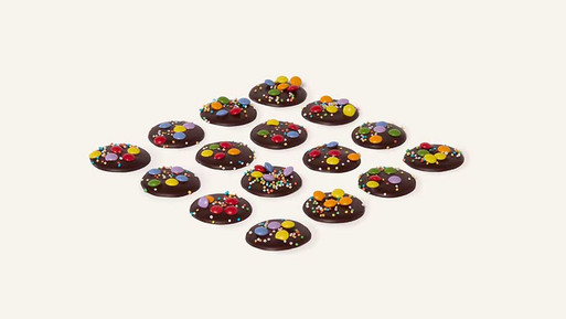 Viele kleine, runde Palets au Chocolat aus Zartbitter Schokolade mit bunten Schokolinsen in Rasteranordnung auf hellem Hintergrund – feine, französische Schokoladenplätzchen mit farbenfrohem, spielerischem Finish aus der Breuninger Confiserie.