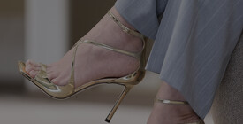 Goldene Jimmy Choo-Sandaletten, getragen zu grauer Hose auf Teppichboden.