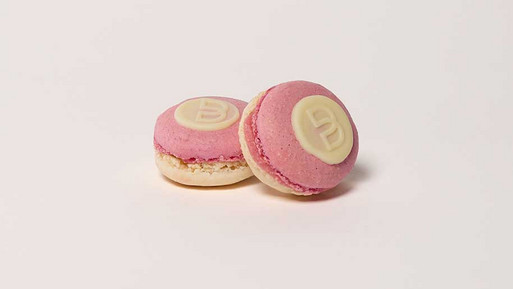 Zwei Strawberry-Cheesecake-Macarons mit rosa und weißer Schale, heller Füllung und einem runden Siegel aus weißer Schokolade mit Breuninger „B“ auf der Oberseite, vor hellem Hintergrund.