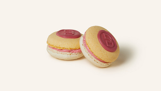 Zwei Pfirsich-Himbeer-Spritz-Macarons mit einer sanft apricotfarbenen Schale, verziert mit einem rosafarbenen Schokoladenmongramm mit dem Breuninger B. Gefüllt mit einer rosa Ganache, vereinen sie die süße Leichtigkeit von Pfirsich und Himbeere mit einer spritzigen Note – ein erfrischender Genuss aus der Breuninger Confiserie.