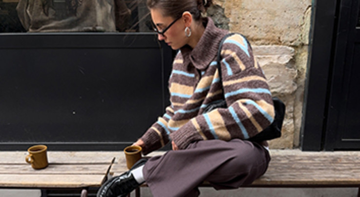Eine Frau die mit zwei Bechern kaffee auf einer Holzbank sitzt. Sie trägt einen gestreiften Pullover, eine braune Hose und schwarze Schuhe. Sie hat braune Haare und eine Sonnenbrillen. 