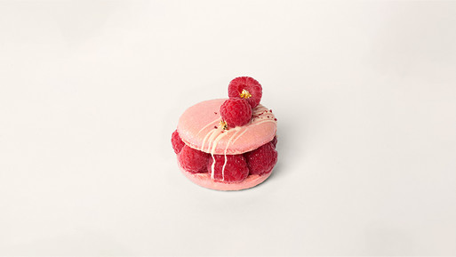 Elegantes Macaron-Törtchen mit zartem Rosaton, gefüllt mit cremiger Ganache und umhüllt von frischen roten Himbeeren, gekrönt mit weiteren Himbeeren und feinen goldenen Perlen – ein süßer Traum aus der Breuninger Confiserie.