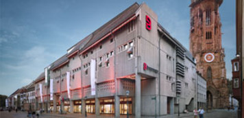 Außenansicht des Breuninger-Hauses in Freiburg bei Abenddämmerung. Das moderne Gebäude mit grauer Fassade trägt das rote Breuninger-Logo. Daneben ist der historische Freiburger Münster mit seinem hohen Turm zu sehen.