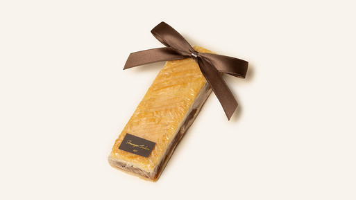 Ein traditioneller Engadiner Nussriegel aus der Breuninger Confiserie, der mit seiner feinen Mischung aus zartem Mürbeteig und einer Füllung aus karamellisierten Walnüssen begeistert. Der Riegel ist sorgfältig in transparente Folie verpackt, wodurch seine köstlichen Schichten sichtbar bleiben und seine handwerkliche Qualität betont wird. Eine dunkelbraune Schleife aus hochwertigem Band rundet die Verpackung ab und verleiht dem Nussriegel eine elegante Note. Das dezente Etikett mit der Aufschrift „Breuninger Confiserie 1951“ unterstreicht die traditionsreiche Herkunft. Ein edler Genuss für besondere Momente oder als liebevolles Geschenk.